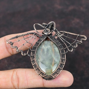 May include: A copper wire wrapped pendant featuring a green gemstone. The wire is woven into a delicate design resembling wings.