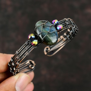 May include: A copper wire bracelet with a woven design and a large oval labradorite gemstone. The bracelet is accented with iridescent glass beads.