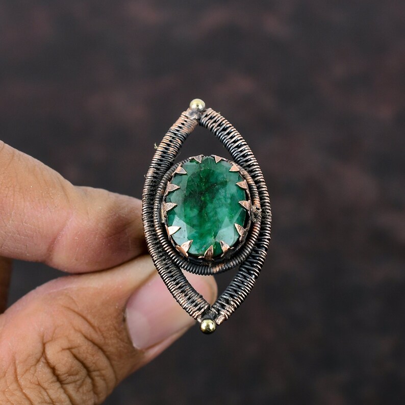 May include: A green gemstone ring with a copper wire setting. The ring is oval shaped and has a wire design that resembles an eye.