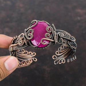 May include: A handcrafted copper cuff bracelet with a large, oval magenta gemstone. The intricate wirework design surrounds the stone and decorates the bracelet's band. The open-ended design allows for easy wear.