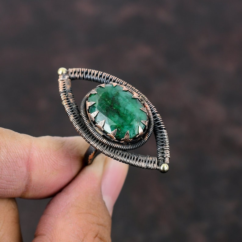 May include: A green gemstone ring with a copper wire design in the shape of an eye. The ring is made of copper wire and has a green gemstone in the center.