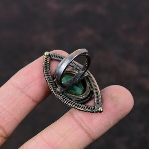 May include: A silver ring with a green gemstone set in a wire-wrapped design. The ring is shaped like an eye and has a gold accent.