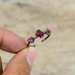 May include: A bronze-colored adjustable ring featuring three raw, vibrant red ruby gemstones. The ring's design is organic, with the gemstones set along a delicate band. The ring is held by a person's fingers.