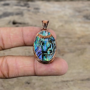 Op de afbeelding: Ovaalvormige hanger met een koperen frame en een kleurrijke abalone schelp inlay. De hanger heeft een koperen beugel om een ​​ketting aan te bevestigen.