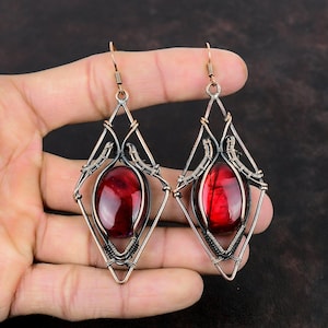 May include: A pair of dangle earrings with a red gemstone set in a copper and black wire frame. The earrings are in the shape of a diamond.