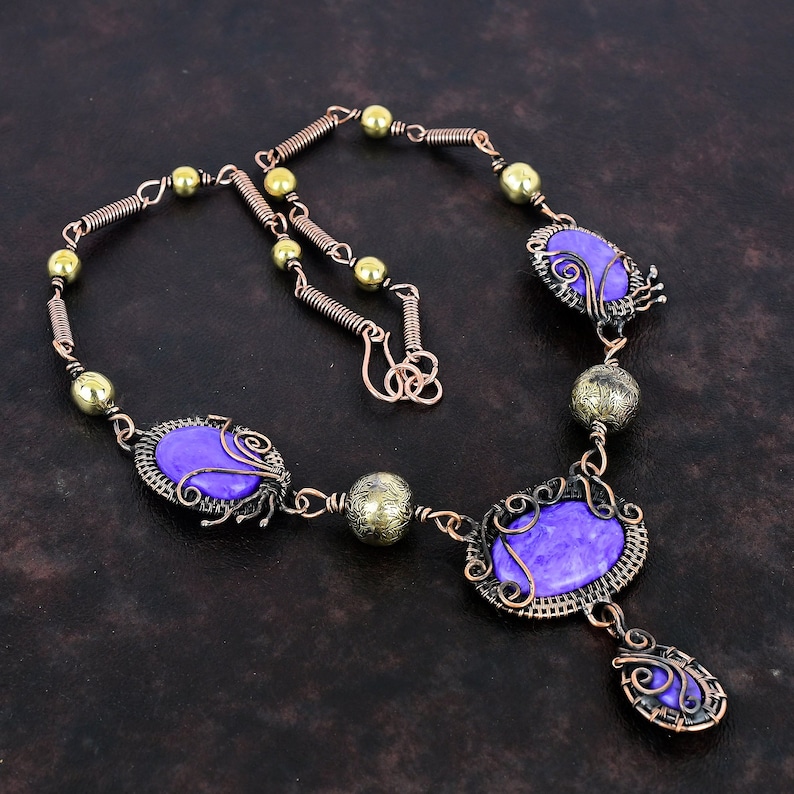 May include: A handcrafted necklace featuring purple oval-shaped stones set in ornate copper wire frames. The necklace includes brass-colored beads and a copper wire chain with a hook clasp. The design has a vintage, bohemian aesthetic.