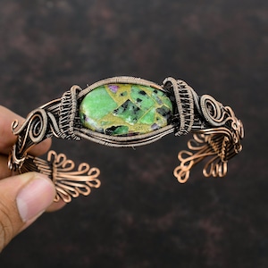 May include: A copper wire wrapped bracelet with a green, pink, and gold gemstone. The bracelet has a delicate, intricate design.