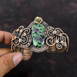 Puede incluir: Brazalete de puño de cobre hecho a mano con una gran piedra ovalada verde y magenta. El brazalete presenta intrincados trabajos de alambre y un pequeño adorno de rosa dorada. El diseño es ornamentado y detallado, con una estética vintage.