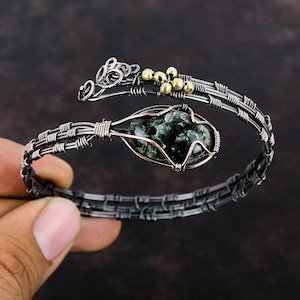 May include: A silver wire wrapped bracelet featuring a green and black stone with gold accents.