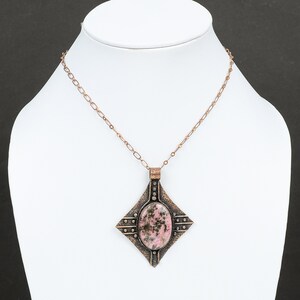 May include: A copper chain necklace with a square pendant featuring a pink and black stone. The pendant has a textured, hammered metal design.