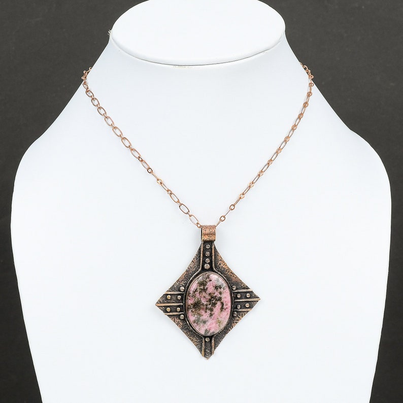 May include: A copper chain necklace with a square pendant featuring a pink and black stone. The pendant has a textured, hammered metal design.