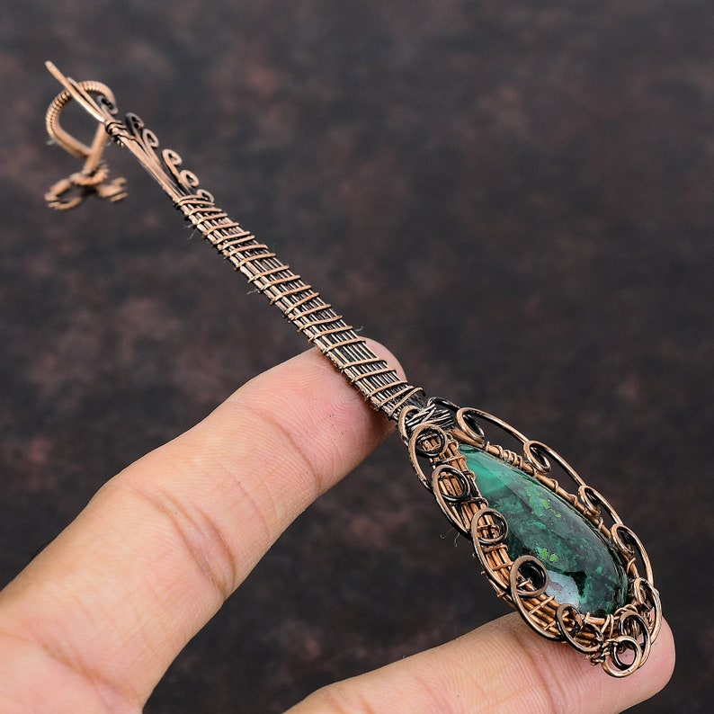 May include: A handcrafted copper pendant with a teardrop-shaped green stone. The pendant features intricate wire wrapping and a decorative bail. The stone is a vibrant green colour, and the copper has a warm, antique finish. The pendant is held by a finger.