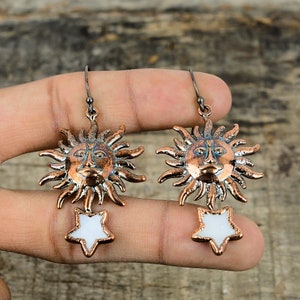 May include: A pair of copper-toned earrings with a sun and star design. The sun features a face and rays, and the star is white with a copper outline.