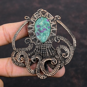 May include: A handcrafted copper pendant in the shape of an octopus. The pendant features a central oval-shaped stone with green and purple hues, encased in a woven copper frame. The octopus design includes detailed wire tentacles.