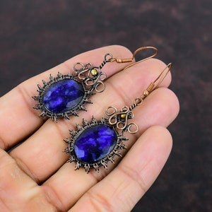 May include: A pair of handcrafted earrings featuring large, oval, deep blue gemstones. The gemstones are set in a dark metal frame with a sunburst design. The earrings have copper-colored wire detailing and ear wires, creating a unique, artistic look.