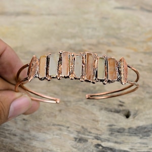 May include: A copper cuff bracelet with a row of rectangular, multi-coloured stones set in the centre. The stones are a mix of white, brown, and pink.
