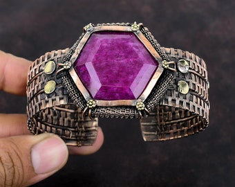May include: A handcrafted copper cuff bracelet featuring a large, faceted, hexagonal magenta gemstone. The bracelet has a woven wire design with decorative brass accents. The cuff is held by a hand.