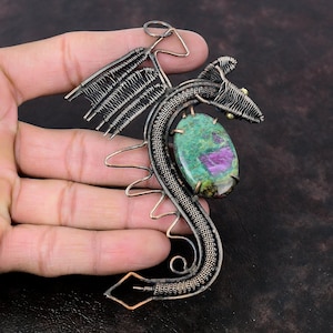 Puede incluir: Un colgante de dragón de alambre de cobre con una piedra preciosa verde y rosa. El dragón está enroscado alrededor de la piedra preciosa, que está engastada en el centro del colgante.