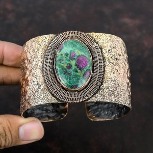 May include: Wide cuff bracelet with a hammered gold-tone finish. The centerpiece features an oval-shaped green and red stone, framed by a woven wire design. The bracelet has an open design and is made of metal.