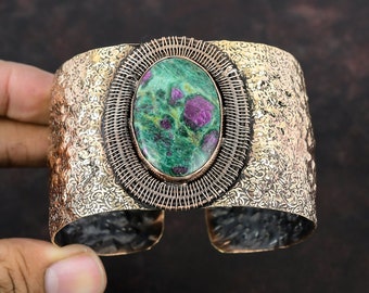 May include: Wide cuff bracelet with a hammered gold-tone finish. The centerpiece features an oval-shaped green and red stone, framed by a woven wire design. The bracelet has an open design and is made of metal.