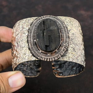 Könnte beinhalten: Ein breites Armband mit strukturiertem Metall-Finish. Das Armband hat einen großen, oval geformten schwarzen Stein, der aufwendig verdrahtet ist. Das Metall hat ein warmes, antikes Aussehen, und die Manschette ist unten offen.