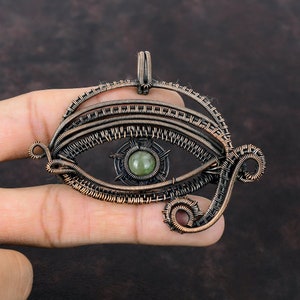 May include: A wire-wrapped pendant shaped like the Eye of Horus, crafted from dark oxidised copper wire. The design features a central round, smooth, pale green gemstone set within a detailed, multi-layered wire frame with intricate coiled accents.