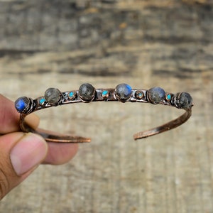 May include: A copper bangle bracelet with a row of six dark gray labradorite stones and smaller turquoise stones between them.