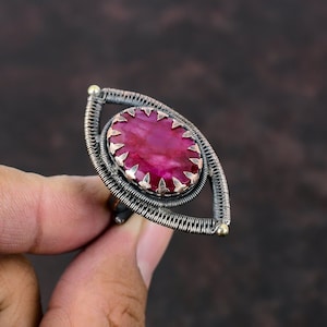 May include: A handcrafted ring with a large, oval, deep pink gemstone set in a wire-wrapped, eye-shaped frame. The ring has a silver and copper-toned metal design with gold accents. The gemstone is approximately 2.5 cm in length.