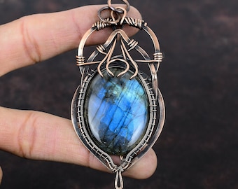 May include: A copper wire wrapped pendant featuring a blue labradorite gemstone. The pendant has a delicate, intricate design with a teardrop shape.