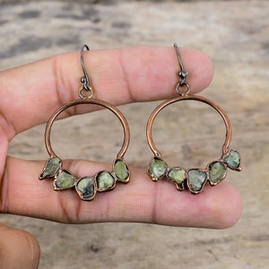 Może przedstawiać: Miedziane kolczyki koła z serią jasnozielonych, surowych kamieni szlachetnych. Kolczyki mają ciemne metalowe haczyki i są prezentowane na jasnobrązowej drewnianej powierzchni.