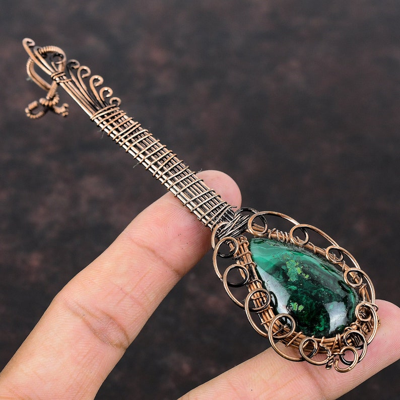 May include: A handcrafted copper pendant with a malachite stone. The pendant features an oval-shaped green malachite stone, intricately wire-wrapped in copper. The pendant's design includes a long, woven handle and decorative swirls.