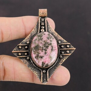 May include: A square-shaped pendant featuring an oval-shaped pink and green gemstone set in a bronze-coloured metal frame. The frame has intricate etched designs and a decorative border.