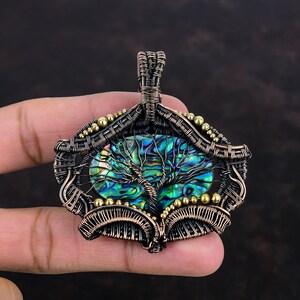 Peut inclure: Un pendentif en fil métallique enveloppé avec une incrustation ovale en nacre d'ormeau irisée. Une silhouette d'arbre en fil de cuivre sombre est superposée sur la coquille. Le cadre est fait de bobines de cuivre et de petites perles dorées.