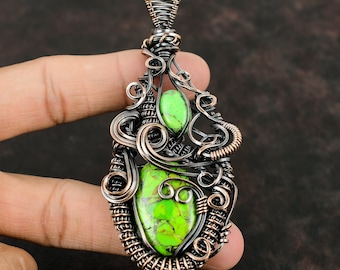 May include: A handcrafted pendant featuring two vibrant green stones set within an intricate wire-wrapped design. The pendant's frame is a mix of copper and dark metal, creating a striking contrast. The pendant is approximately 2 inches long.