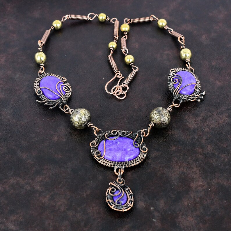 May include: A handcrafted necklace featuring purple oval and round stones, accented with copper wire and brass beads. The necklace has a unique, artistic design with intricate wirework and a vintage aesthetic.