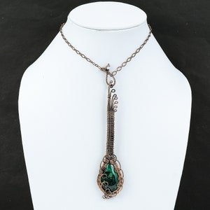May include: A handcrafted copper wire pendant necklace with a green stone. The pendant has an elongated design with intricate wire wrapping and a delicate chain. The necklace is displayed on a white mannequin bust against a black background. The pendant is approximately 8 cm long.