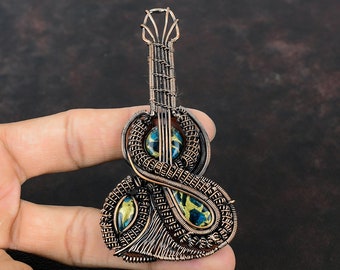 May include: A copper wire wrapped guitar pendant with blue and gold glass inlay. The pendant is held in a hand.