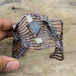May include: A copper wire cuff bracelet with a wavy design and embedded semi-precious stones. The stones are various shades of blue, white, and gray.
