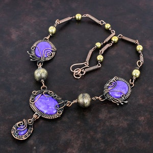 May include: A handcrafted necklace featuring purple gemstone cabochons set in ornate copper wire frames. The necklace includes decorative copper wire links, brass beads, and a hook clasp. The design is intricate and artistic, showcasing a bohemian style.