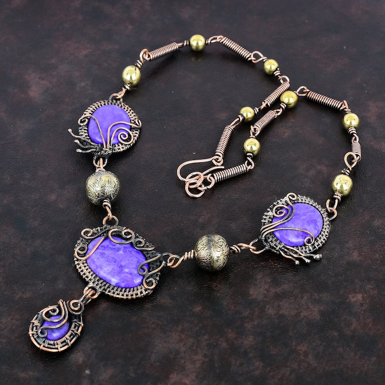 May include: A handcrafted necklace featuring purple gemstone cabochons set in ornate copper wire frames. The necklace includes decorative copper wire links, brass beads, and a hook clasp. The design is intricate and artistic, showcasing a bohemian style.