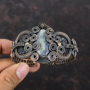 May include: A silver and copper wire wrapped bracelet with a gray and white gemstone in the center. The bracelet is decorated with intricate wire spirals.