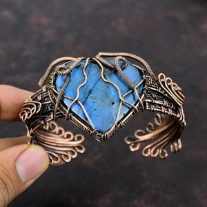 May include: A copper wire wrapped bracelet with a large blue labradorite gemstone in the center. The gemstone is set in a heart shape and the bracelet is decorated with intricate wirework.