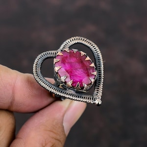 May include: A handcrafted ring with a large, faceted, oval-cut ruby gemstone. The ruby is set in a copper-coloured bezel with star-shaped accents. The ring's band is a heart-shaped design made of woven silver-coloured wire.