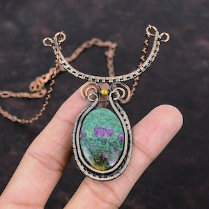 May include: A handcrafted copper wire pendant necklace. The pendant features a large, oval-shaped green stone with red inclusions, encased in a woven copper wire frame. The necklace has a delicate chain and a decorative copper wire design.