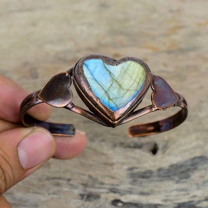 May include: A copper bracelet with a heart-shaped labradorite gemstone. The bracelet is open and has two smaller heart-shaped copper accents on either side of the gemstone.