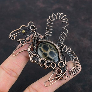 May include: A copper wire sculpture of a squirrel with a black and white agate stone in the center. The squirrel's tail is curled up and the stone is surrounded by intricate wire details.