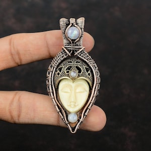 May include: A copper wire wrapped pendant featuring a white carved face with closed eyes and a crown. The pendant is accented with iridescent moonstone gems.