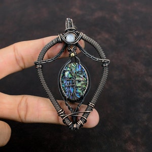 Op de afbeelding: Handgemaakte hanger met een draadgewikkeld ontwerp. De hanger toont een abalone schelp met een levensboom ontwerp, geaccentueerd door een maansteen en goudkleurige kralen. Het draadwerk is donkergrijs en koperkleurig, wat een uniek, artistiek stuk creëert.