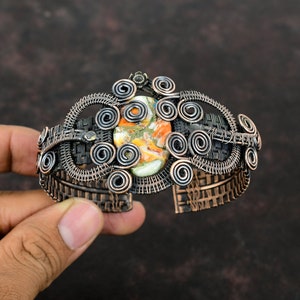 Könnte beinhalten: Ein Armreif aus dunkel oxidiertem Kupferdraht mit komplexen Spiralmustern und einem geflochtenen Metallband. In der Mitte befindet sich ein ovaler Stein mit marmorierten Mustern in den Farben Orange, Gelb, Weiß und Hellgrün.