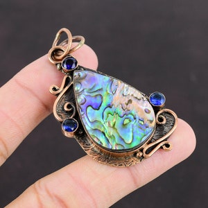 Op de afbeelding: Een traanvormige hanger met een koperen setting en een grote, iriserende abalone-schelp inlay. Drie kleine blauwe edelstenen zijn in het koper rond de schelp gezet.